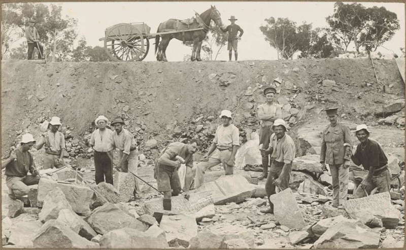 Sepia photo of men in hats carrying tools standing in rubble in a pit. In the background is a horse and cart led by another man in a hat. 