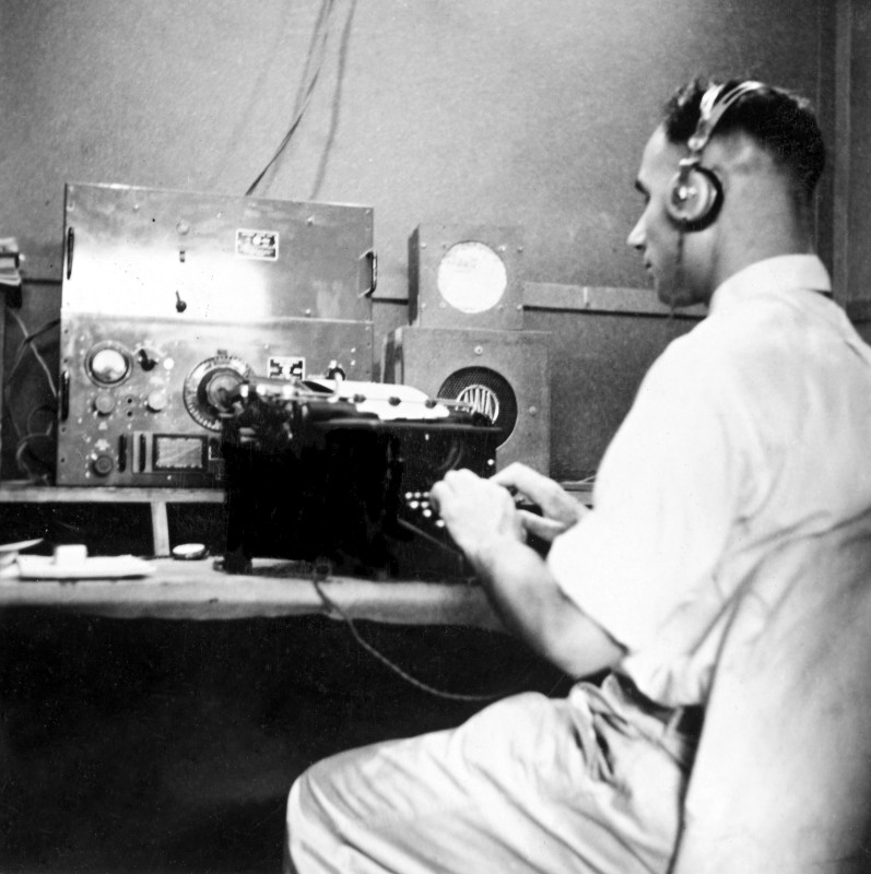 Black and white photograph of a man wearing headphones and using a typewriter inside a radio operations centre.