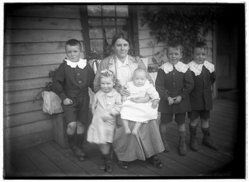 Black and white photograph of a woman seated on verandah of weatherboard house. She has a baby on her knee and a little girl leaning against her other knee. Three little boys stand beside her.