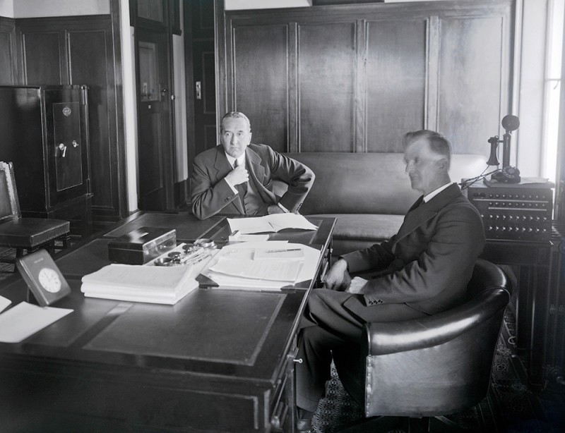 Black and white photograph of two men sitting at a large desk. Both wear suits and have paperwork in front of them.