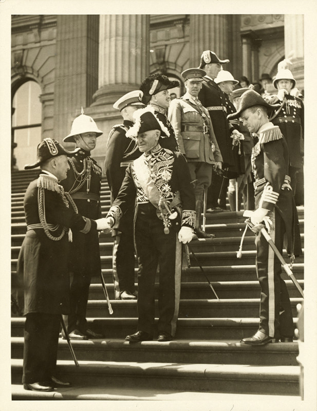 Several men wear formal military uniforms and hats and stand in a row down stairs at the front of a building. A man wearing a feathered hat, formal decorated suit, sash and gloves shakes hands with one of the men in the line.