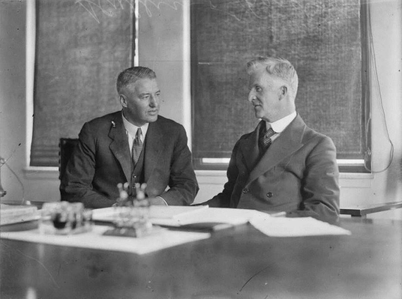 Black and white photo of two men wearing suits and ties sitting side-by-side at a desk. There are papers laid out on the table in front of them. 