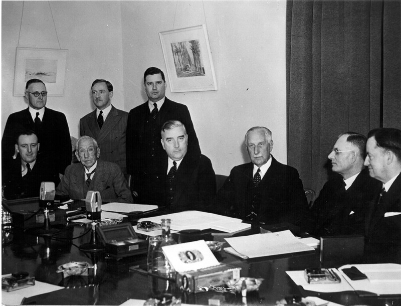 Six men seated at a meeting table with papers in front of them. Three men stand behind them.