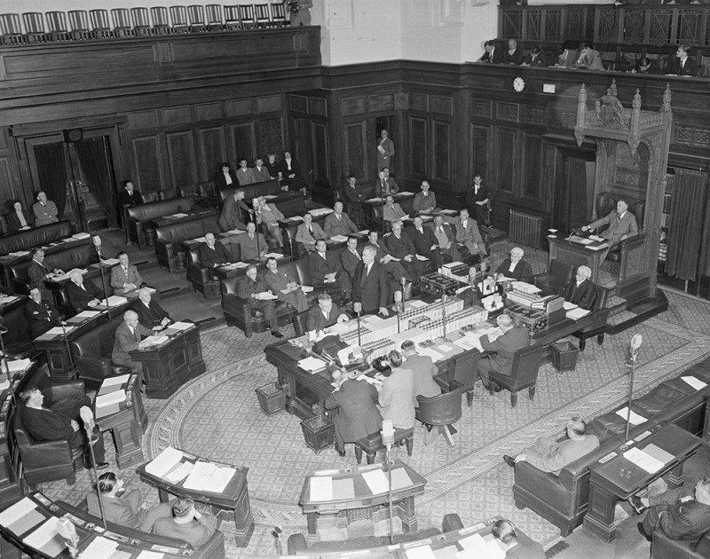 A large room where many men in suits are sitting in a U-shaped seating arrangement. A man stands to speak to the other men. 