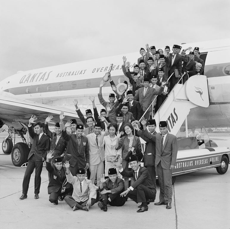 A group of around 35 people stand on the steps leading into an aircraft. They are smiling and waving at the camera.