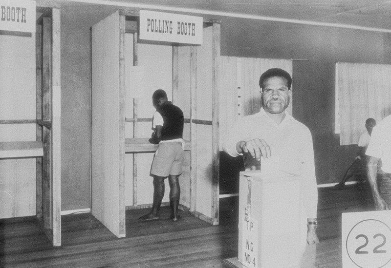 A black and white photo showing a man placing a ballot paper into a slit in the top of a metal box. In the background, another man stands with his back turned inside a small polling booth filling in a ballot paper.