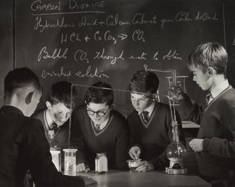 A black and white photograph showing five schoolboys standing around a table in a science classroom conducting an experiment.