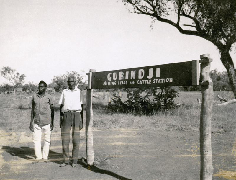 Two Aboriginal men stand next to a large sign in the outback.