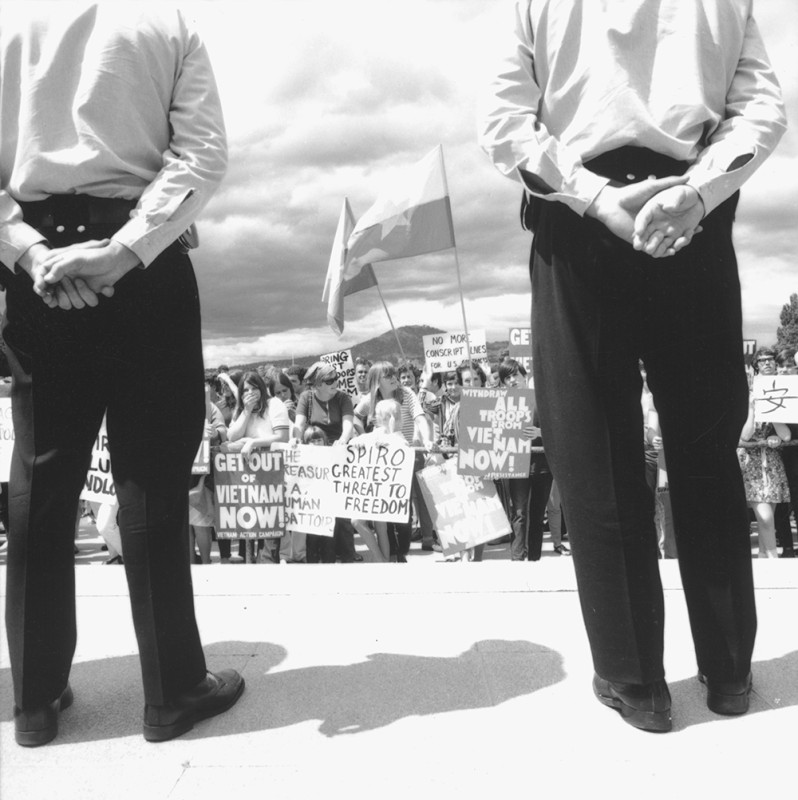 A group of people holding signs. The backs of two men wearing uniforms can be seen watching the crowd.