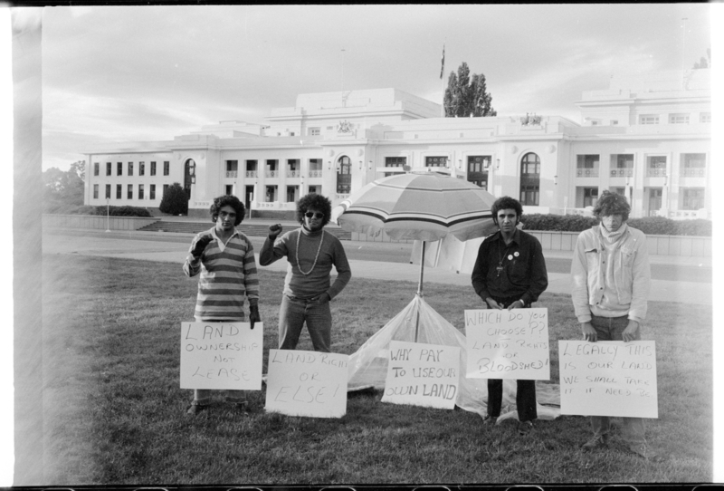 4 men standing near a beach umbrella and holding signs in front of a large building.