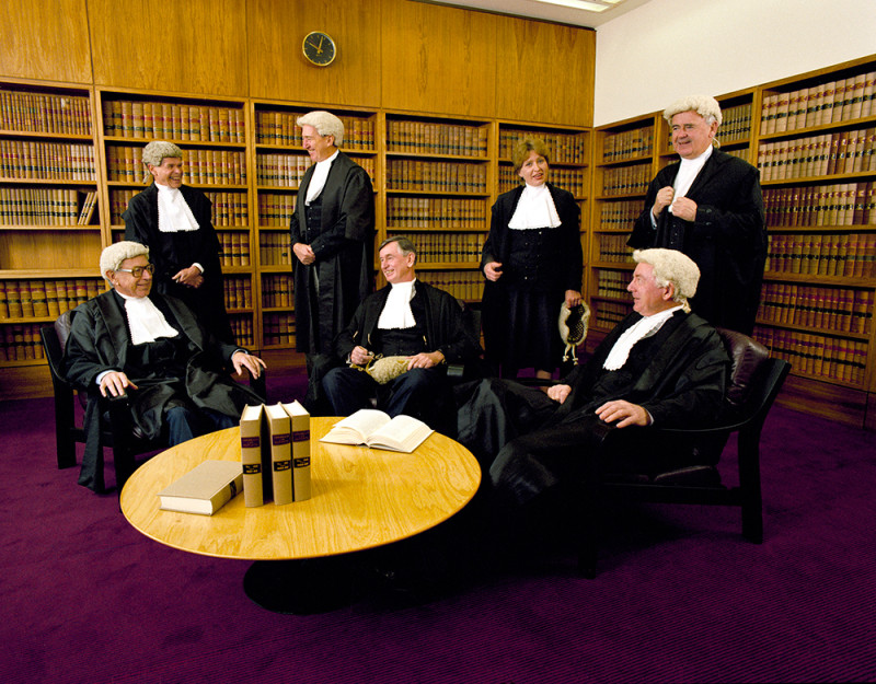  6 men and 1 woman in a room with books on shelves around the walls. They all wear black robes. 5 of the men wear white court-room wigs.