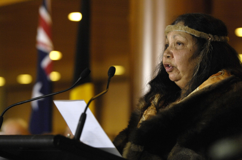 A woman standing at a podium delivers a speech. She wears a possum skin cloak and headband.