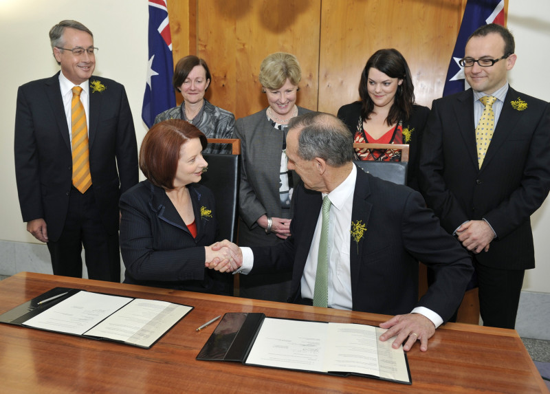 Two people sit at a desk and turn to shake hands. 5 people stand behind them watching in front of Australian flags. Signed papers and pens are on the desk.