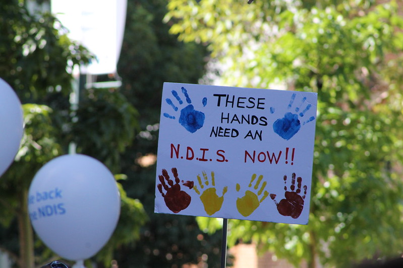 A homemade placard with 6 handprints and text that says ‘These hands need an N.D.I.S. now!!’.