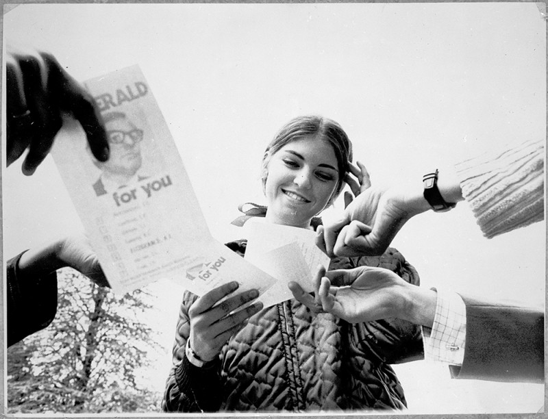 A young woman is handed how-to-vote flyers. She smiles while looking at the flyers and has one hand on her head.