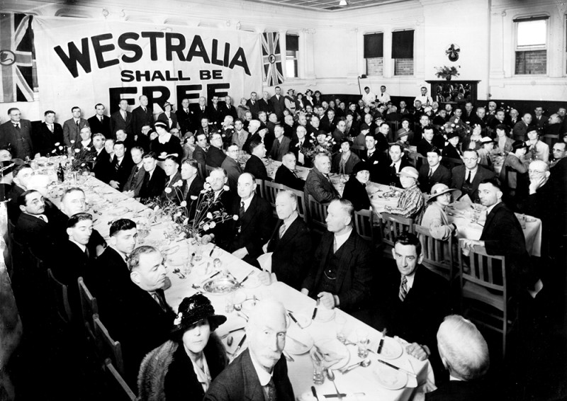 A large group of people sit around 4 long tables. In the background is a flag with the text ‘Westralia Shall Be Free.’ 