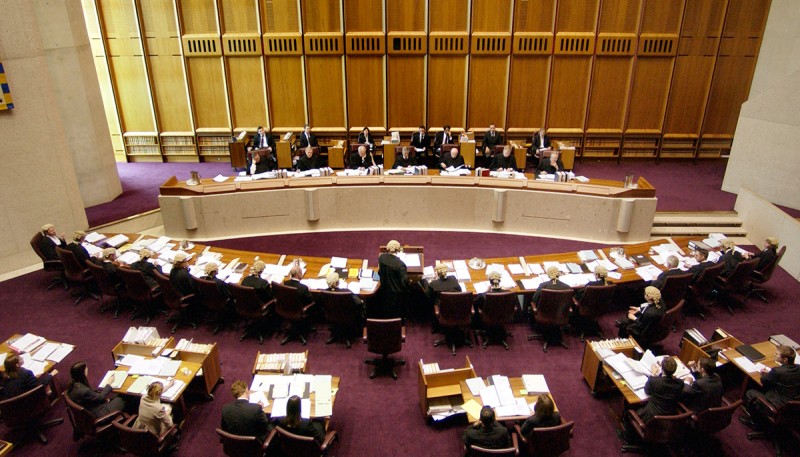 The Australian High Court in session. Judges wearing black robes sit at a long table. They are facing lawyers sitting at another table. The lawyers wear old-fashioned wigs.