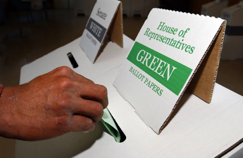 A white hand is putting a green piece of paper in a white box labelled 'House of Representatives/GREEN/Ballot Papers'