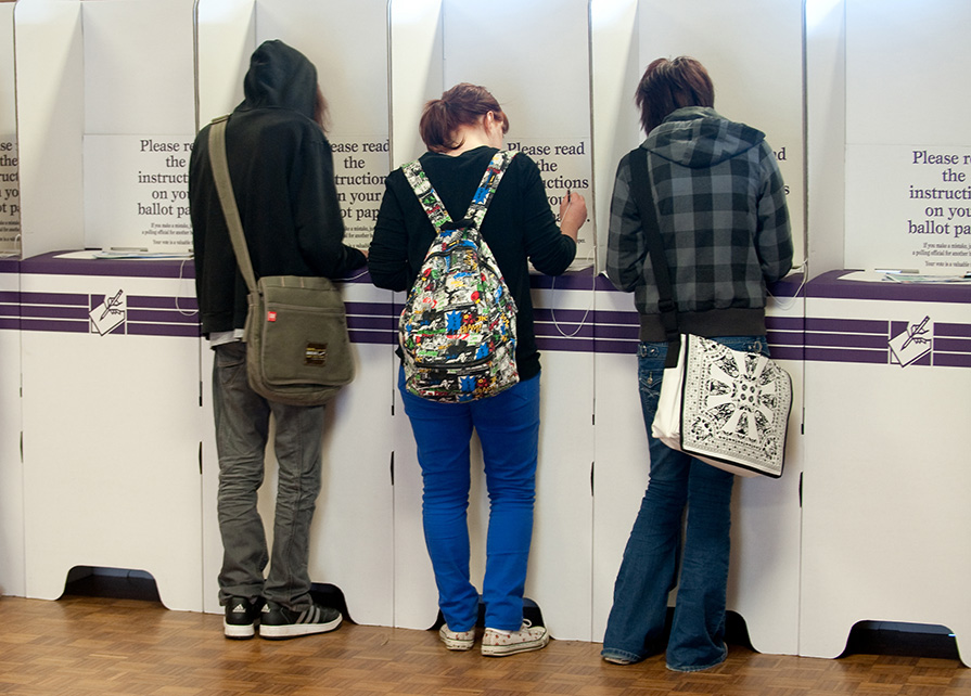 A photo of 3 people voting at a row of purple and white voting booths, with their backs to the camera.