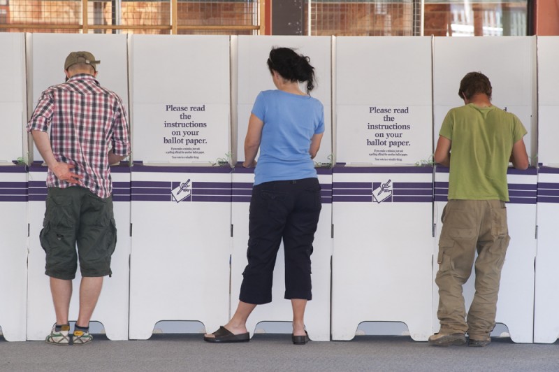 Three people dressed in casual clothes stand at white ballot boxes. They have their back to us