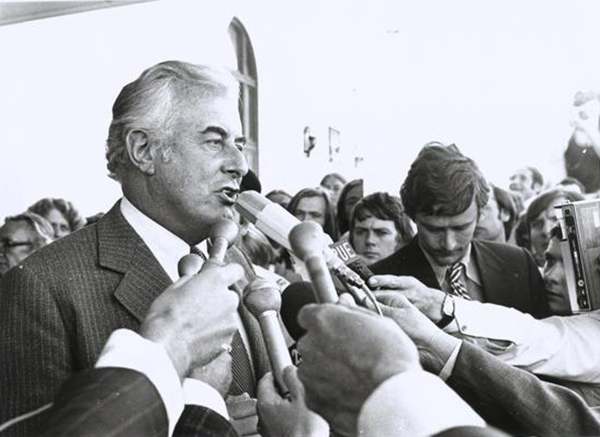 Prime Minister Gough Whitlam surrounded by the press. He is talking into microphones and is surrounded by people.