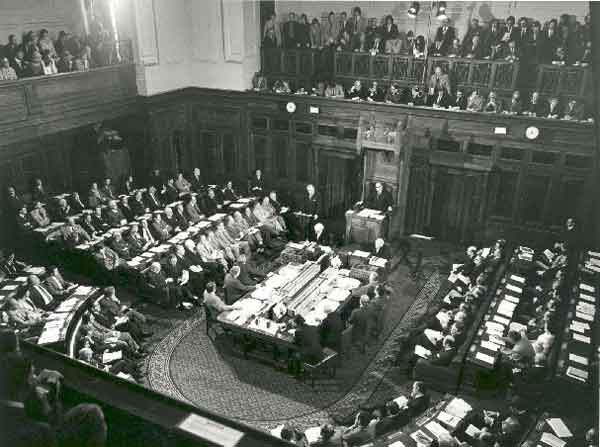 Black and white photo of the House of Representatives chamber with horse shoe shaped seating and all seats full.