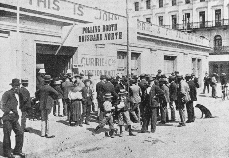 A black and white photograph of people standing outside a polling place.