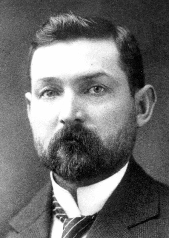 A black and white portrait of a man wearing a dark suit, white shirt and dark tie. He has dark hair and a short beard and moustache. 