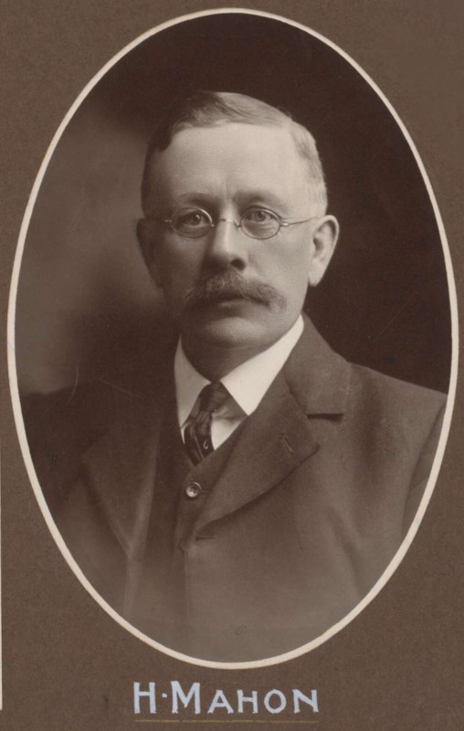 A sepia-toned photograph of a middle aged man with a moustache and round wire-rimmed glasses. He is wearing a suit with a thin tie.
