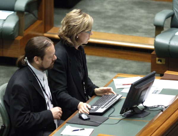 A man and a woman dressed in dark clothes sit at a small table. On the table is a computer and papers. The woman holds the keyboard while looking to her left.