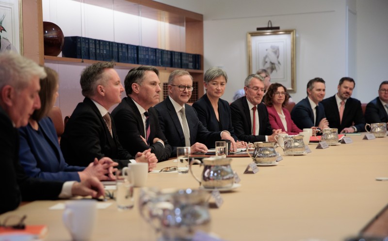 A group of people having a meeting sit both sides of a wooden table. The men and women look towards the camera.