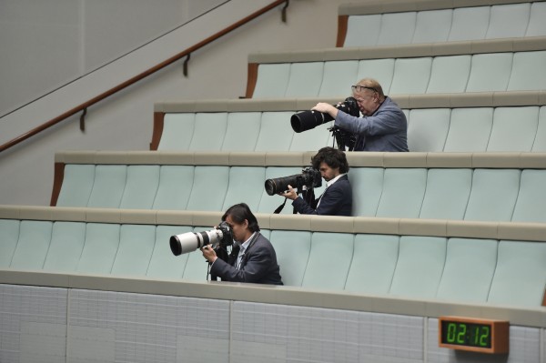 Three photographers have their eyes to their cameras. They hold the long zoom lenses. They are seated in teired rows of green seats.
