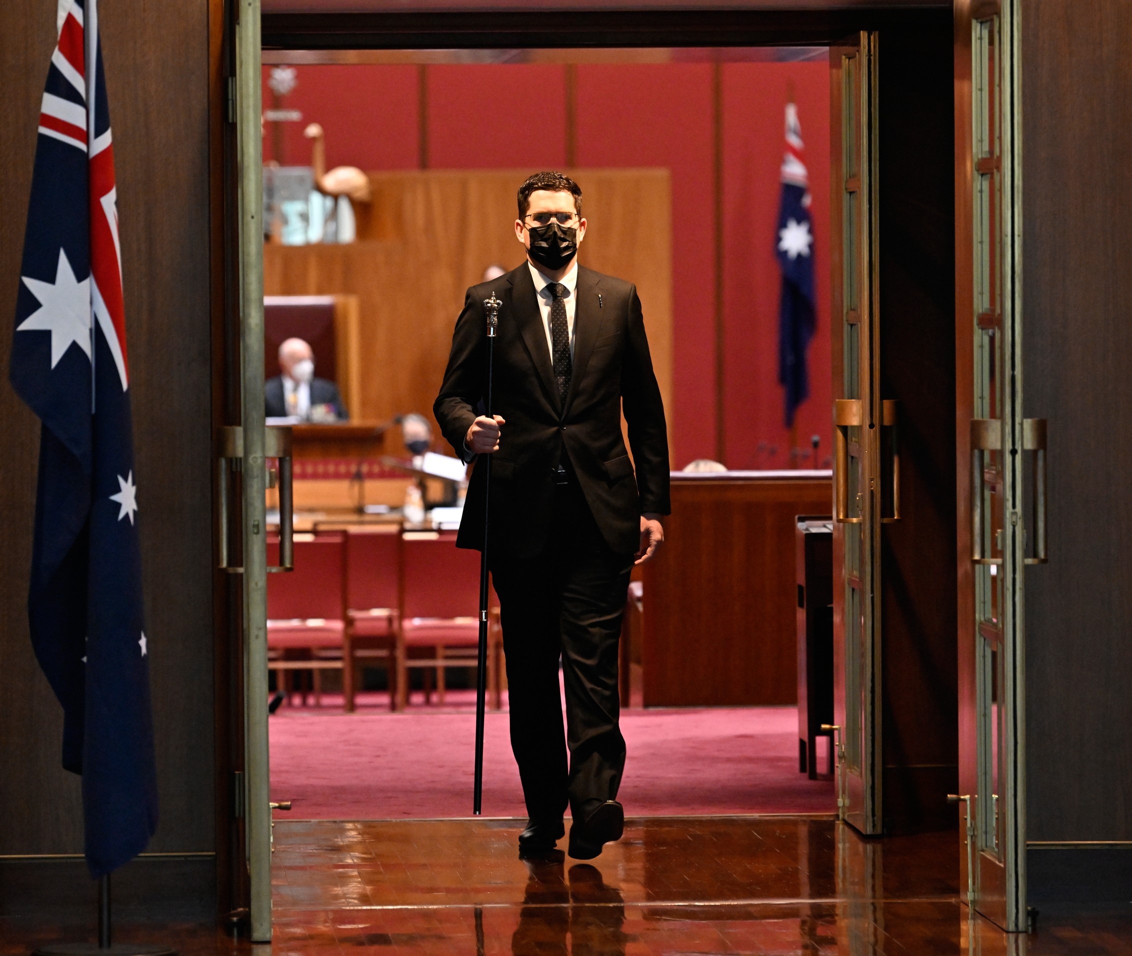 A man wearing suit and holding a black staff with a silver crown is walking through an open doorway. Behind him is a red room (the Senate).
