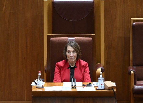 A middle-aged woman dressed in a red jacket sits in a high-backed dark red chair. Behind heris a wooden screen, On the desk infront of her are papers and a bottle of hand sanitiser.