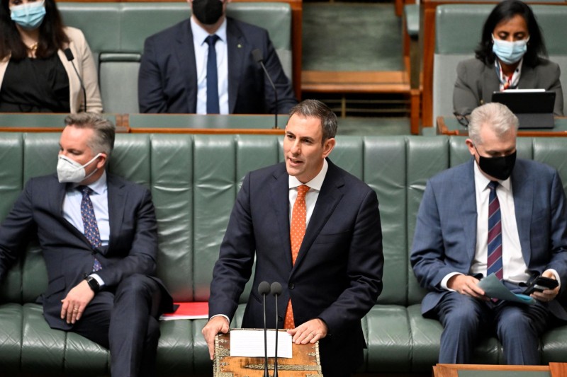 A white man in a suit looks to the left while his left hand rests on a piece of paper on a large box. There is a microphone in front of him. Behind people sit on a green bench