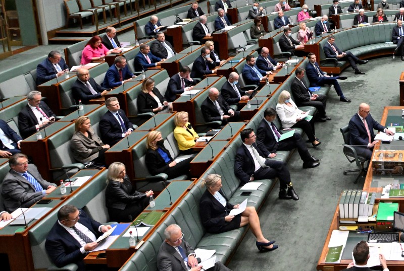 In a room with green seats and carpet there is a large table where the Leader of the Opposition sits. There are 5 rows of seats behind him occupied by members of the Opposition.