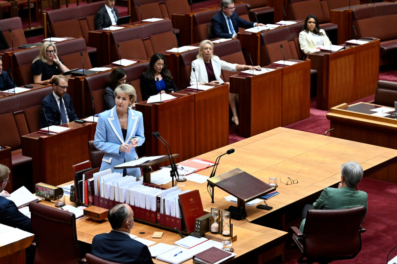 A man in a suit stands at a table and is speaking in a red room. A woman and a man in dark suits also sit at the table.