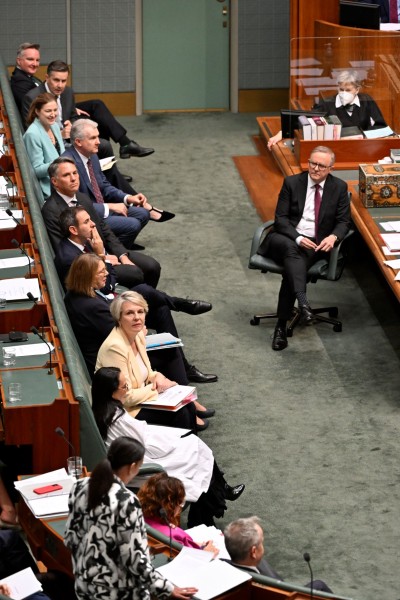 In a room with green carpet and leather seats, men and women sit along a bench. Some are turned to look over their right shoulder to listen to a women standing to give a speech.