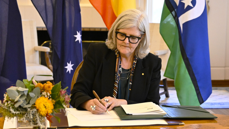 A woman signs a document at desk in front of Australian flags.