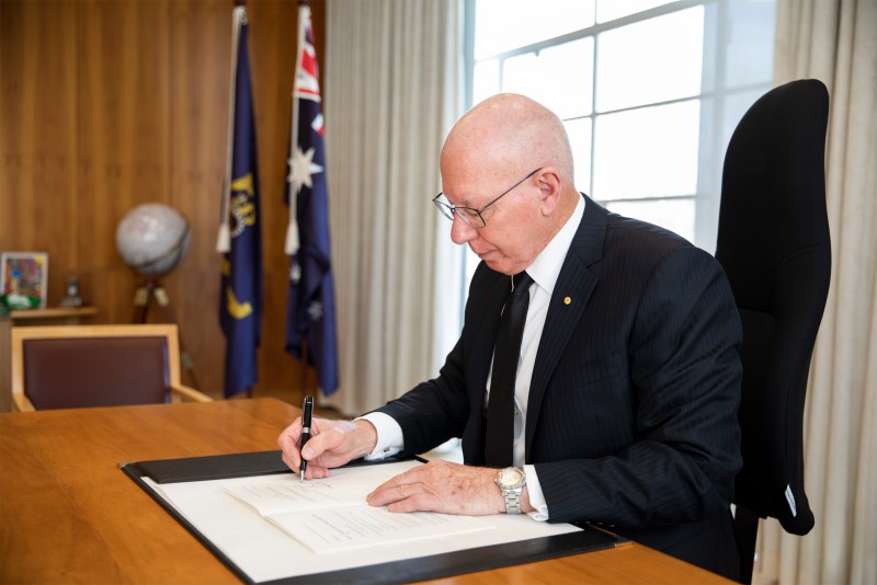 The Governor-General sitting at a desk signing a bill. 