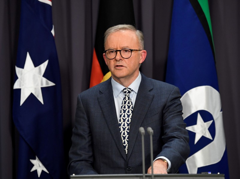 A man in a suit speaking into microphones at a lectern. Behind him is an Australian flag, an Aboriginal flag and a Torres Strait Islander flag.