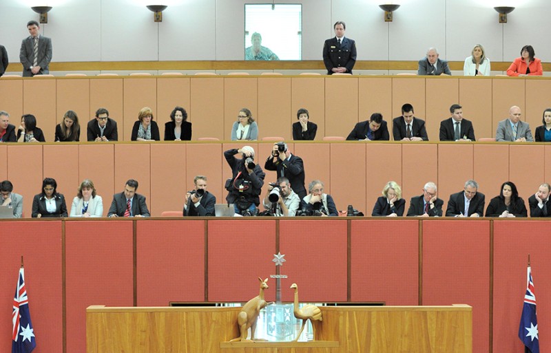 Media with cameras and notebooks sit in tiered seating above a carved wooden Commonwealth coat of arms in the Australian Senate.