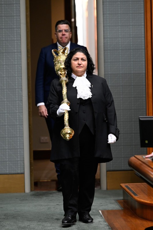 A woman wearing a black suit with a white tie and white gloves is walking into a green room. She carries a large gold mace with the crown over his right shoulder. A man in a suit is walking in behind her.