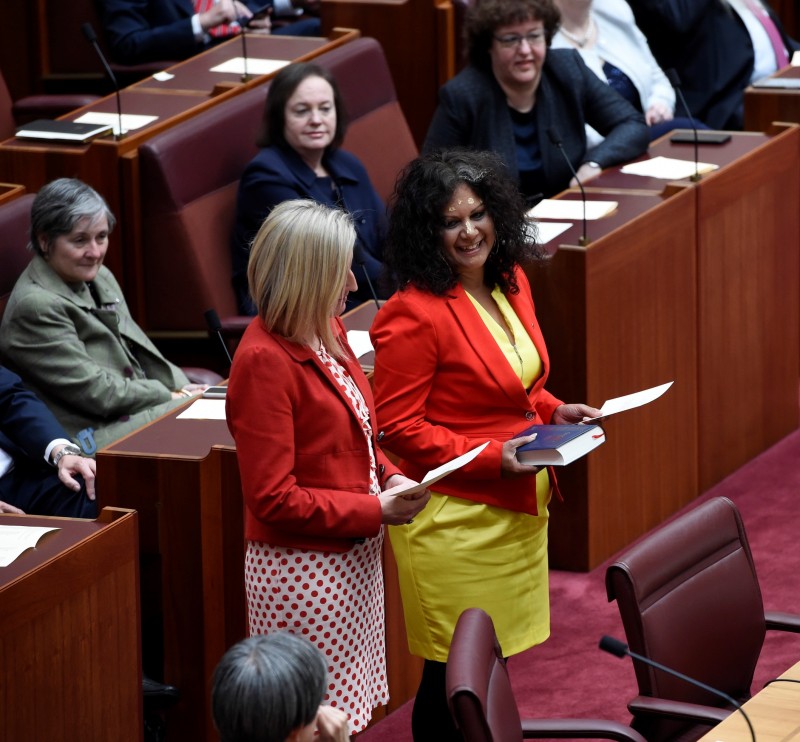Two women dressed in red stand holding pieces of paper. Behind them are people sitting at wooden desks watching. 
