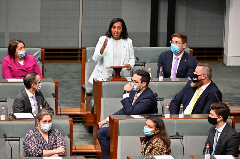 A member of the House of Representatives stands at a desk giving a speech. Other members sit at rows of desks watching and listening to her.