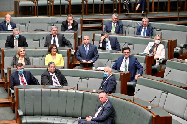 A man wearing a blue suit and tie stands at a desk with his hands on a lecturn. Other men in suits and a couple of women sit listening at thier desks. The chairs and carpet are green.