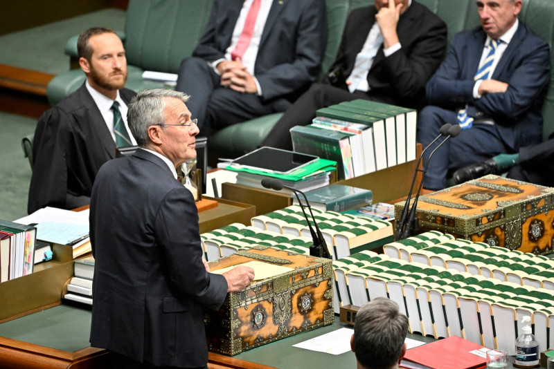 A man in a suit stands at a large wooden table. In the centre are rows of large books lined up with the spines up. The books are bound in green and cream binding.