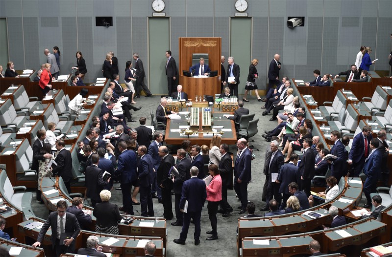 The green House of Representatives. Members are standing and walking across the room, or are already sitting down on the benches.