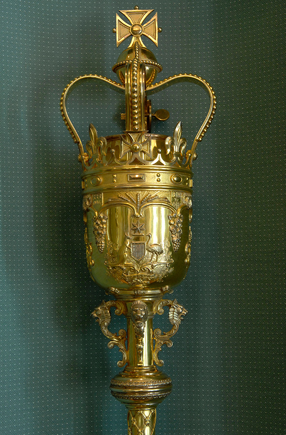 A detailed view of the gold Mace clearly showing the crown with its emblems and etchings. The Mace is standing upright. 