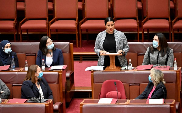 A woman in a grey jacket is standing at a desk in a red room. She is speaking. Other women sit on benches around her, listening to her speak.